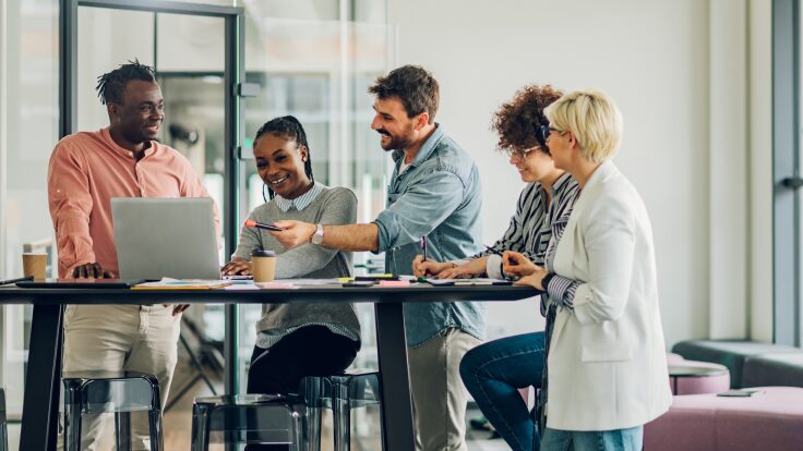 Five people engaged in discussion around a laptop at a high table in a modern office setting, exploring the potential of AI Video Generators to enhance their project's multimedia capabilities.