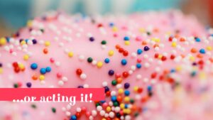 Close-up of a biteable pink frothed doughnut sprinkled with colorful candy beads, with a blurred background and text overlay saying "...or acting it!".