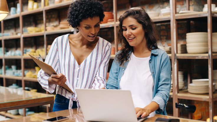 Two women in a pottery shop look at a laptop screen, discussing AI video generators as one holds a clipboard. Shelves filled with pottery grace the background, blending tradition with technology in their creative collaboration.
