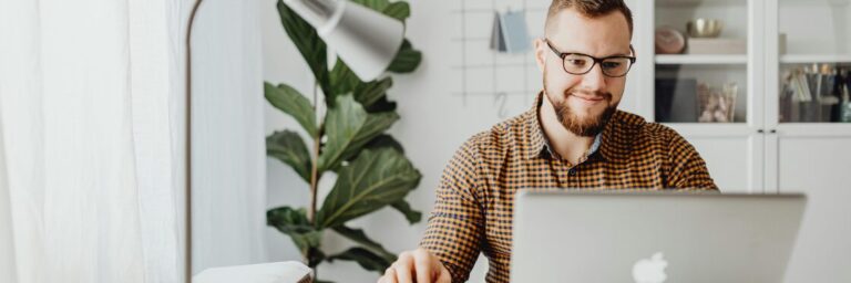 A person with glasses and a beard is sitting at a desk, smiling while using a laptop. A lamp and plant are in the background.