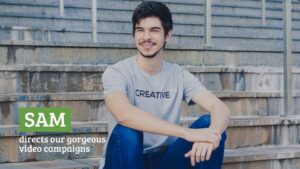 A young man with a gentle smile, wearing a "creative" t-shirt, sitting on outdoor stairs with text overlay "Sam directs our gorgeous Biteable video campaigns.