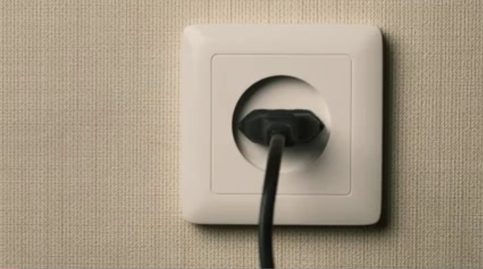 A black electrical plug is partially inserted into a white wall outlet on a beige textured wall.