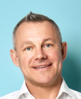 Middle-aged man with a mohawk hairstyle smiling against a turquoise background, wearing a white shirt.