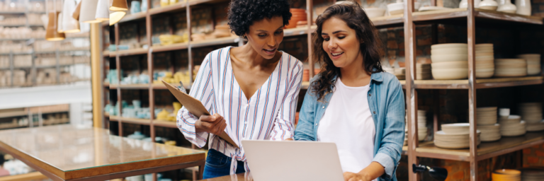 Two women, one holding a clipboard and the other a laptop, discuss work in a pottery shop surrounded by shelves of ceramic goods.