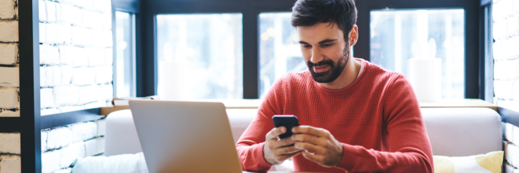 Man in red sweater using smartphone with laptop on table in a modern room with large windows.