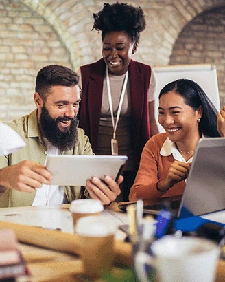 Three colleagues sit at a desk and smile while exploring an online video maker on a tablet, with laptops and coffee cups in front of them in an office setting.